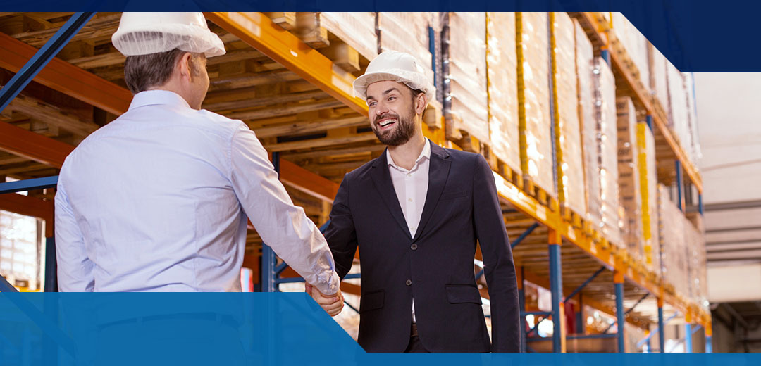 Two men in a warehouse shake hand while wearing hard hats and business clothes